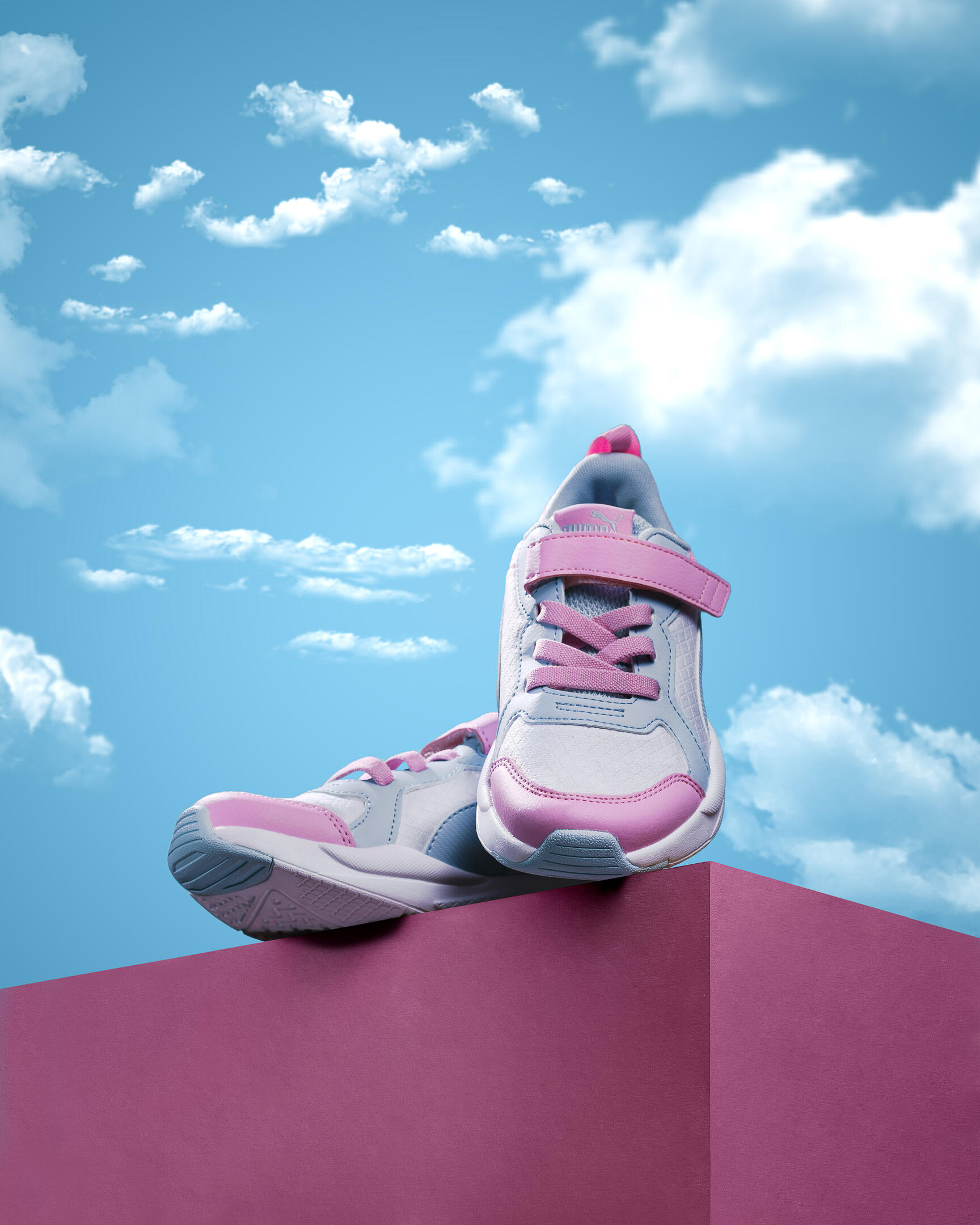 PUMA sneakers on pink cube. On a bright pink cube are PUMA sneakers. Sneakers in pink, white and blue. The color of the sneakers is underlined by the color of the surrounding space in the frame. In the background is a bright blue sky with clouds. A bright sun shines on the sneakers on the left, and a shadow is located below the sneakers, which once again emphasizes the presence of bright light in the frame. 