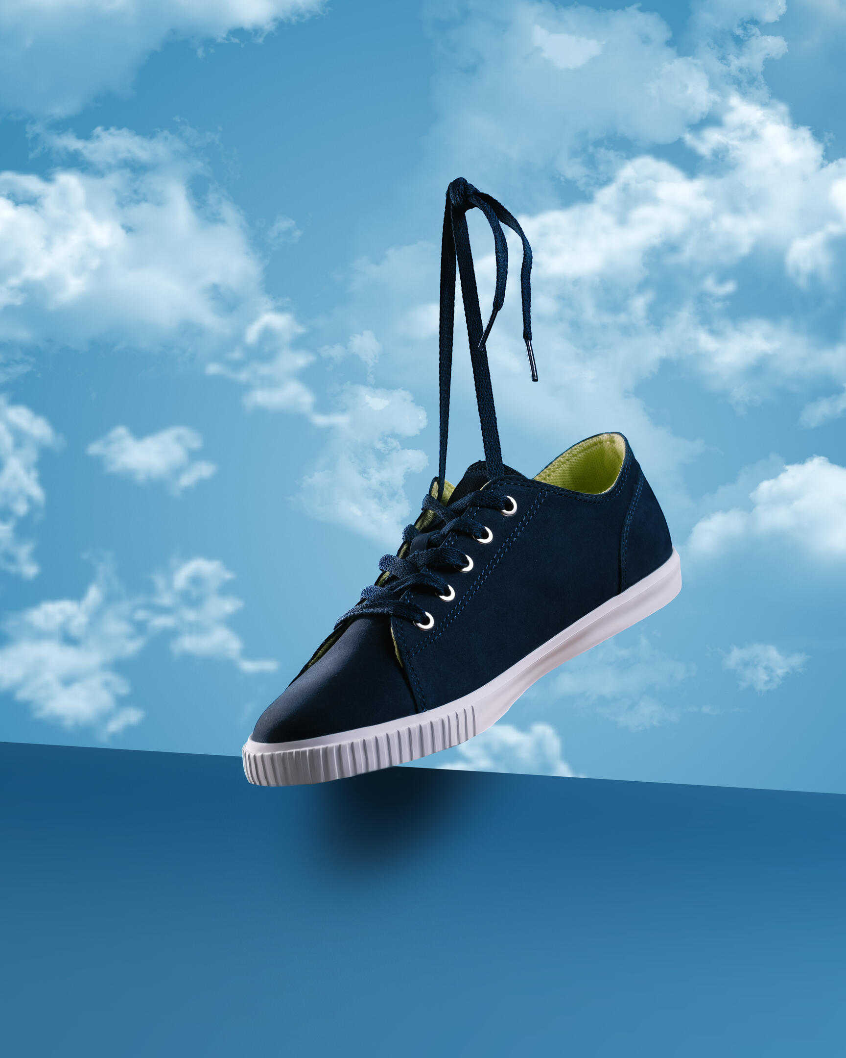 Sneakers - Hero view. Blue canvas sneakers hover in the air and hover over a blue base. The sneakers cast a shadow on the blue stand. In the background, you can see a bright sky with clouds.