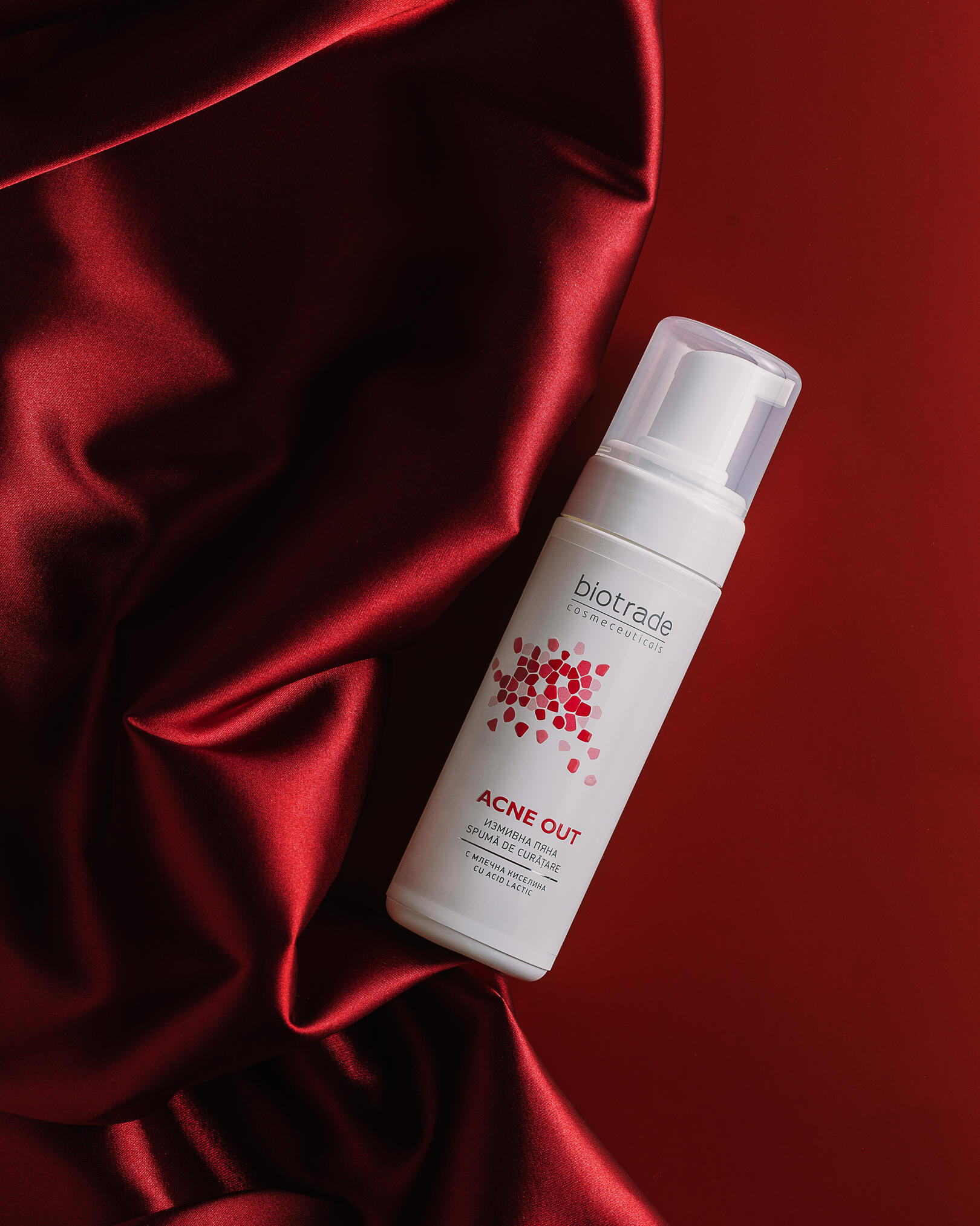  Biotrade Acne Out facial cleanser.  On a red background lies a jar of Biotrade Acne Out facial cleanser. To the left of it is a red fabric - repeating in its color - the color of the pattern on the packaging of the foam. The fabric is shimmery and shiny.