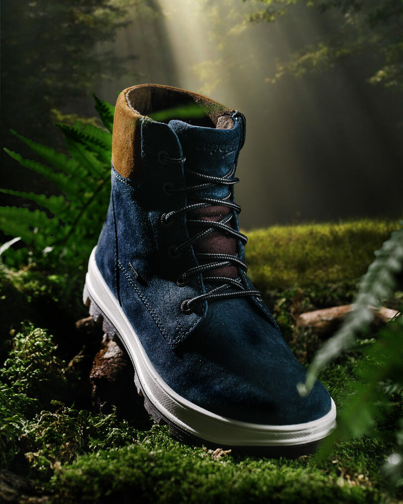Blue suede Superfit boot in a forest. Blue suede Superfit boot with brown toes and laces stand on sunlit moss. The greenery is illuminated by bright sunlight breaking through the foliage of trees.
In the background, rays of sunlight are visible, which pass through the canopy of trees.