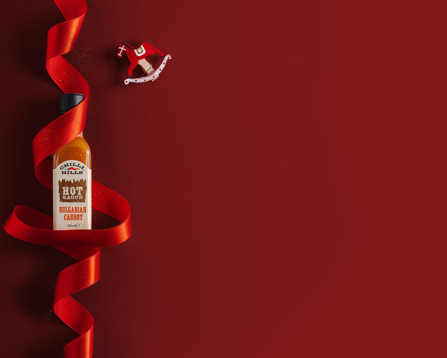 Chilli Hills Bulgarian carrot hot sauce. Christmas mood. On a red background lies a jar of Chilli Hills Bulgarian carrot hot sauce.
A red satin ribbon is wrapped around the jar and sequins are scattered. To the right of the jar, a little above is a clothespin in the shape of a red horse. The satin ribbon lies so that it resembles the shape of a Christmas tree. The entire right side of the picture is free and can be filled with advertising inscriptions.