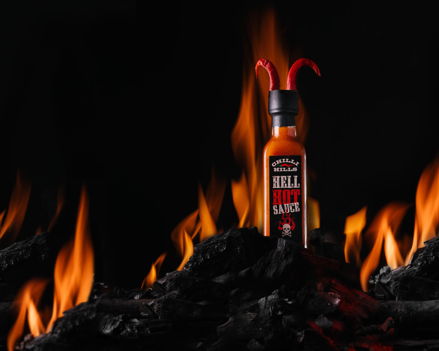 Chilly Hills Hell Hot Sauce на огън. Дървата вече са запалени. Оставяме само въглища... Върху тези изгорени въглища има кутия лют сос Chilly Hills Hell Hot. Тя остана цяла и невредима. На капака на буркана има рогчета от пипер. Пламъкът все още гори зад бутилката. Някои въглища са толкова горещи, че все още са червени.