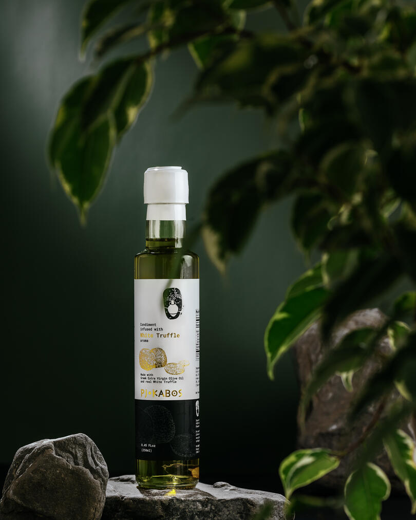 Greek olive oil from PJ KABOS. A bottle of white truffle-scented Greek olive oil from PJ KABOS sits on a gray stone in the sun. The sun shines through the bottle and we see the greenish color of the oil, as well as pieces of truffle at the bottom of the bottle. The foreground is blocked by a bush of greenery. In the background are the same stones.