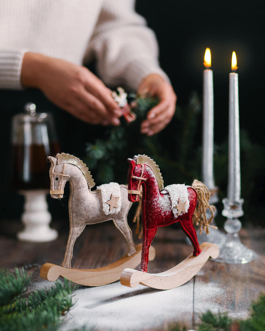 На масата има декоративни играчки Новогодишни люлеещи се коне. На масата има декоративни играчки Новогодишни люлеещи се коне. Жена на фона на рамката украсява клони на коледно дърво. Свещи горят. Нова година е скоро.