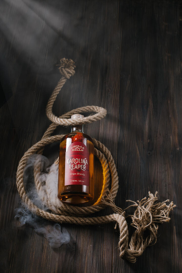 Chilli hills Rakia A bottle of Carolina Reaper Chilli Hills rakia lies on a wooden table. To the left, light enters through a narrow window. The bottle lies on a rope - giving the viewer a reference to the ship theme. Smoke spreads near the bottle - emphasizing the fact that the contents are very hot and spicy.