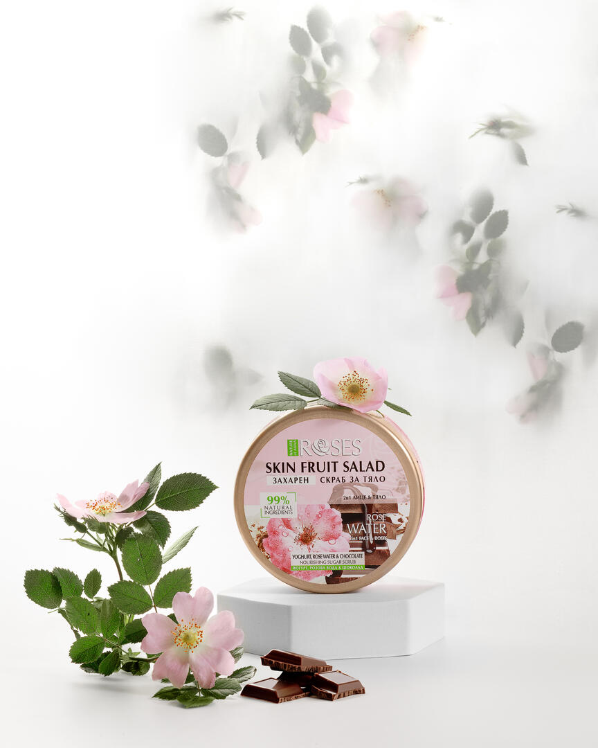 Skin fruit salad scrub for body from Nature of Agiva. 2in1 SUGAR SCRUB yogurt, rose water, and dark chocolate. The scrub stands on a hexagon-shaped stand. Rosehip flowers are visible in the background, which is visible through a light background. Rosehip also lies near the scrub.