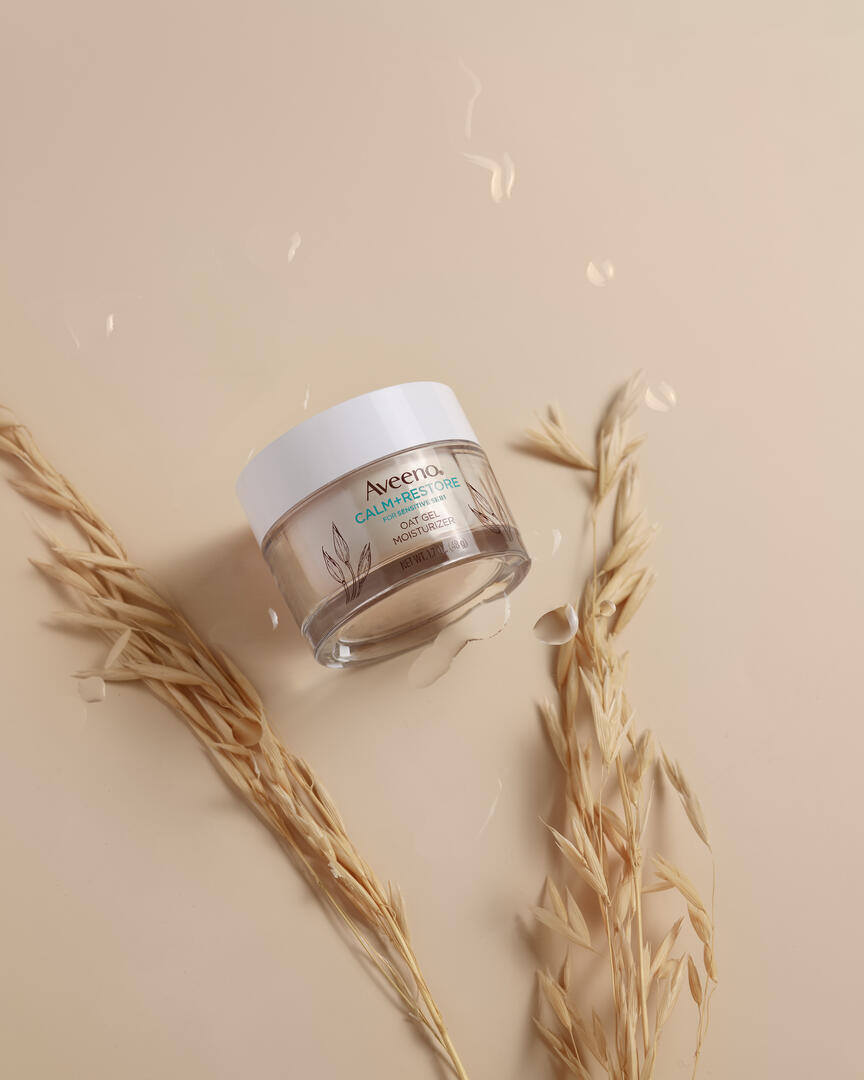 Aveeno oat gel moisturizer Oats lie on a beige background. Near it lies a jar of cream. Drops of water are visible above the jar, emphasizing the cream's moisturizing properties.