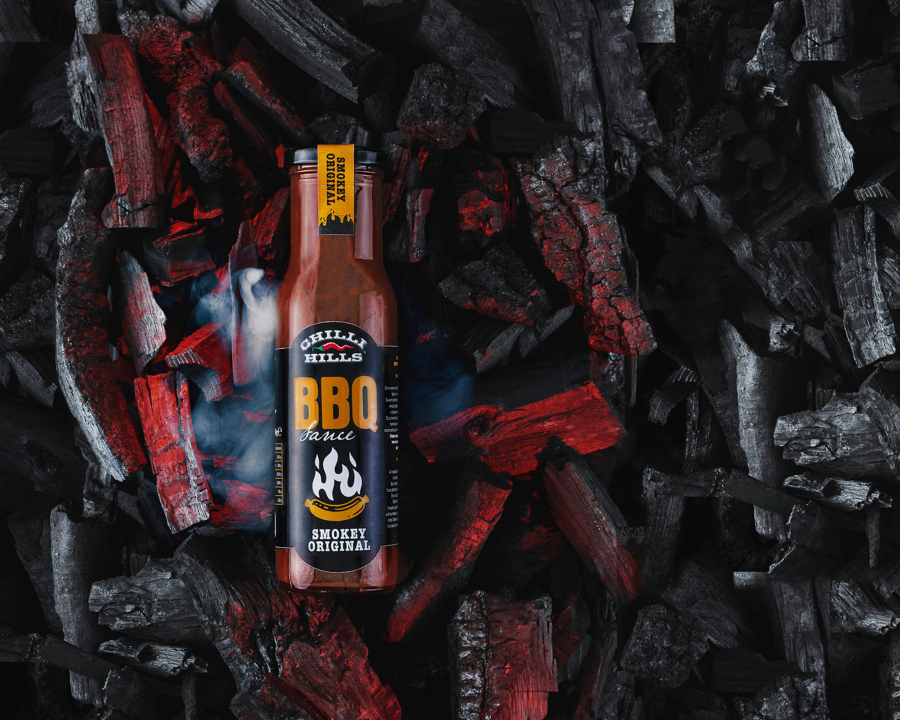 Ketchup Chilli Hills - BBQ Smokey Original. Charcoal surrounds a bottle with ketchup. There is a red glow around imitating the fire and heat.