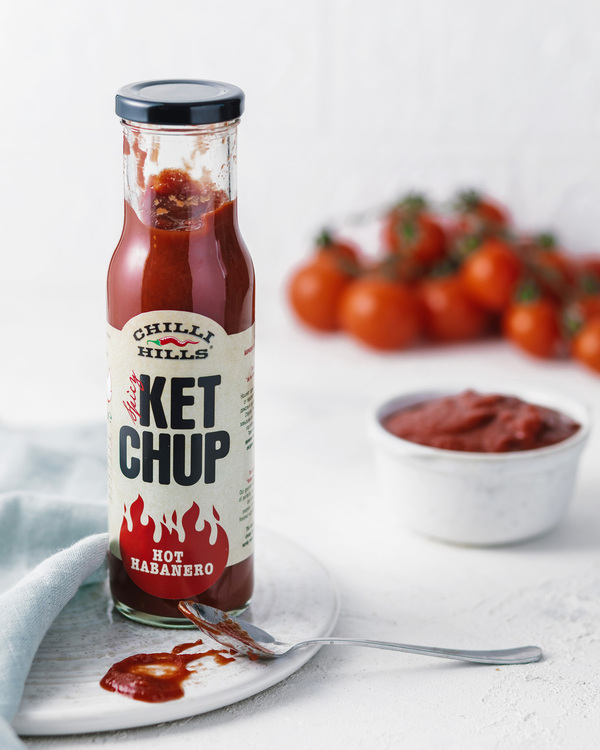 Ketchup Chilli Hills - Hot Habanero. Ketchup Chilli Hills - Hot Habanero. A bottle is in the center. There is a spoon with ketchup in front of it. Fresh tomatoes and a bowl with ketchup are behind.
