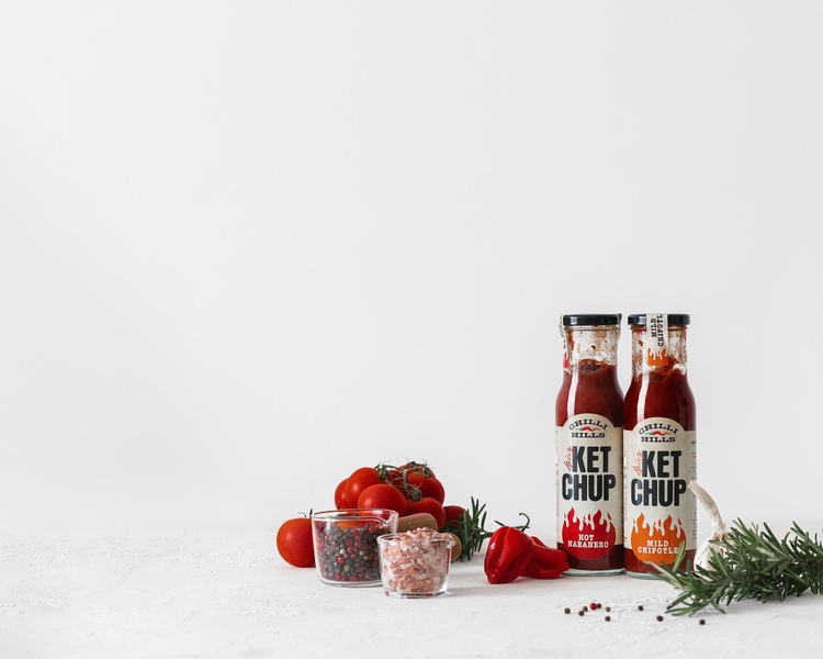 Ketchup Chilli Hills - Mild Chipotle and Hot Habanero. Bottles with ketchup are on a white background surface. Tomatoes, Himalayan pink salt, garlic, black pepper, red pepper, habanero pepper, herbs, and spices surround the bottles.