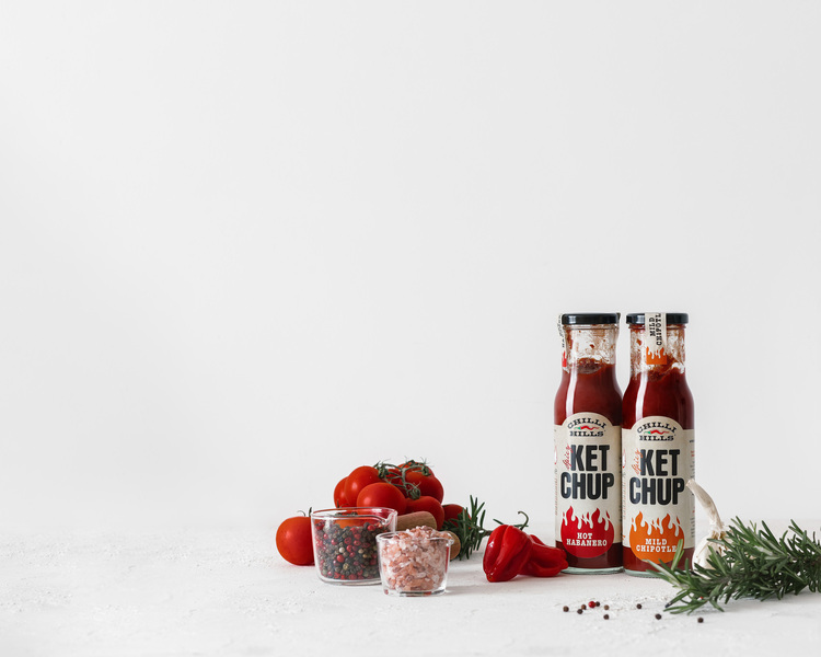 Ketchup Chilli Hills - Mild Chipotle and Hot Habanero. Ketchup Chilli Hills - Mild Chipotle and Hot Habanero. Bottles with ketchup are on a white background surface. Tomatoes, Himalayan pink salt, garlic, black pepper, red pepper, habanero pepper, herbs, and spices surround the bottles.