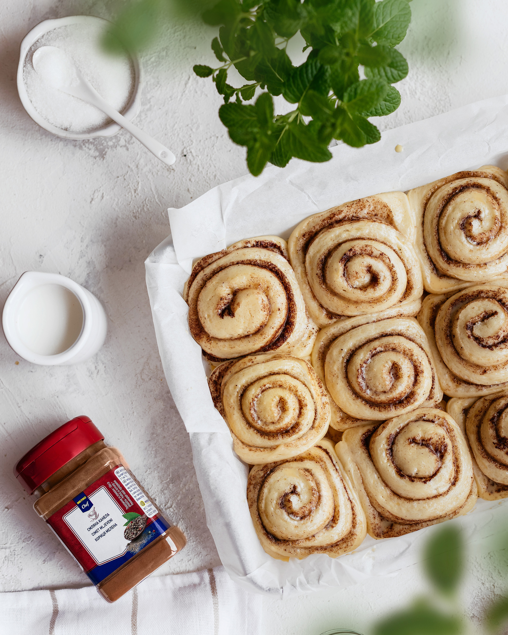 Metro Chef - Ground cinnamon and Cinnabons. Cinnabons (cinnamon rolls) are on a white table with a pack of cinnamon. There is a jar with milk or sour cream, mint, and sugar.