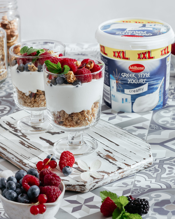 Breakfast in sunny morning with yogurt, and crunches. Breakfast in sunny morning with yogurt, and crunches. There are two transparent tall glasses on the table, they contain crunches, the next layer is yogurt, and berries complete the composition. Berries and fresh mint leaves lie in front of the glasses. In the background are jars of nuts and crunches. To the right of the cups is Milbon Greek Style cream yogurt.