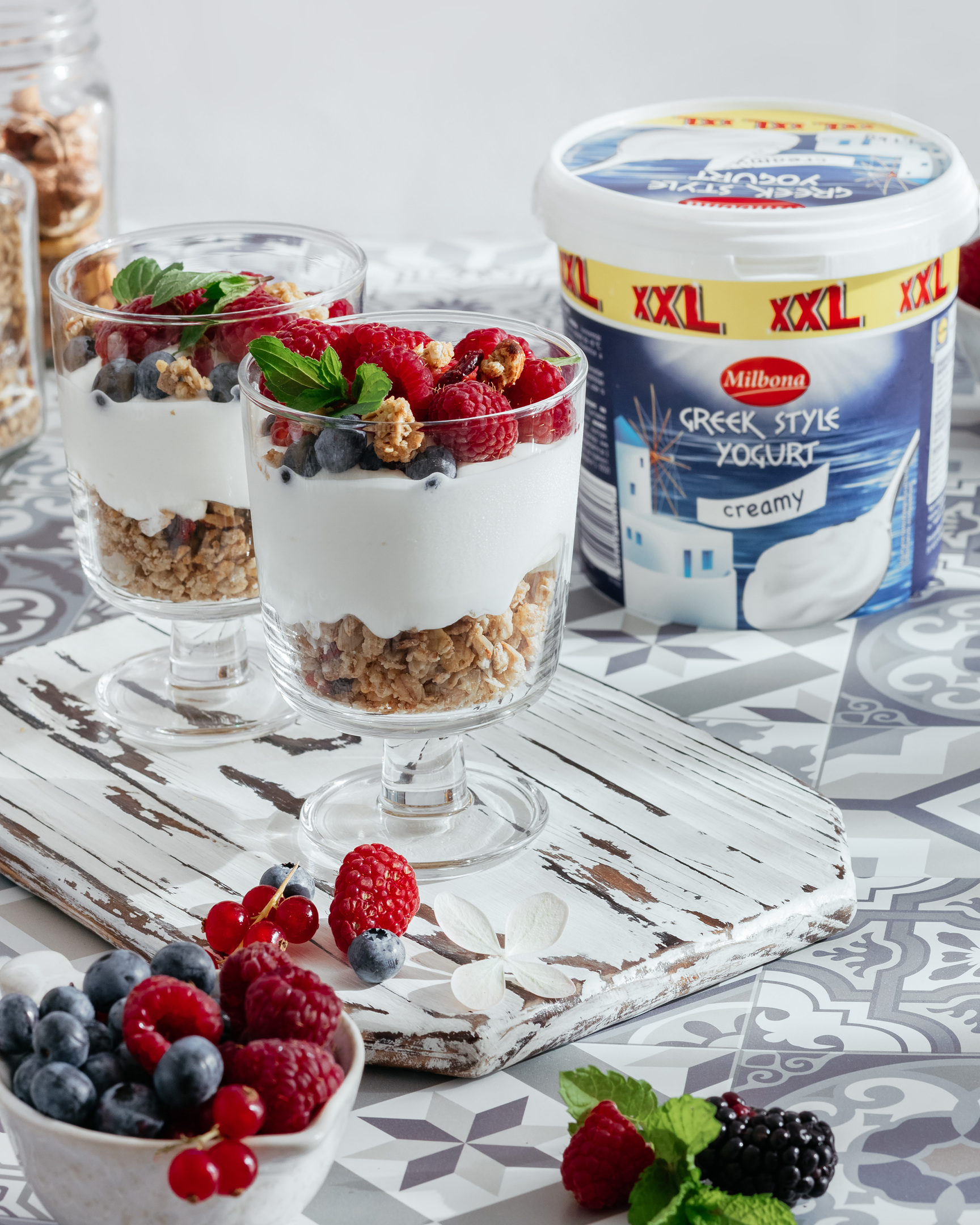 Breakfast in sunny morning with yogurt, and crunches. There are two transparent tall glasses on the table, they contain crunches, the next layer is yogurt, and berries complete the composition. Berries and fresh mint leaves lie in front of the glasses. In the background are jars of nuts and crunches. To the right of the cups is Milbon Greek Style cream yogurt.