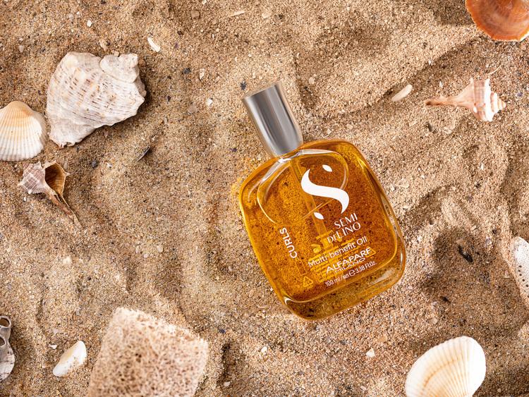 Alfaparf Semi DiLino Oil on Sand. Alfaparf Semi DiLino Oil on Sand. A jar of ALFAPARF Milano Oil for curly hair lies on the sand. Around the jar are various types of shells. The sun shines on the shells, and they cast a shadow on the sand.
