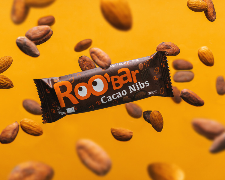Roobar - Cacao Nibs. Roobar is flying surrounded by cacao beans on yellow background.