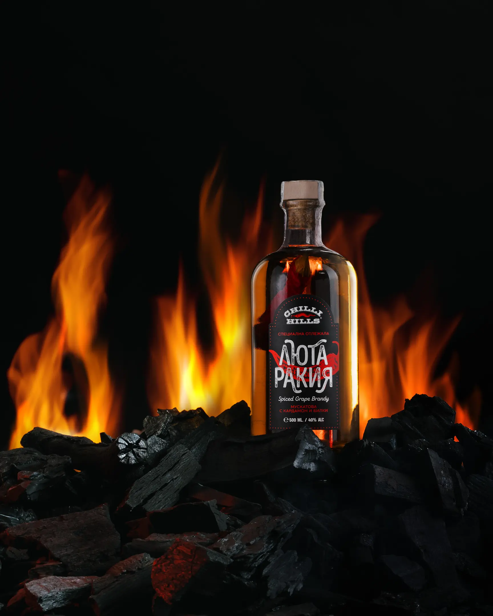 Final Photo. A bottle of Chilli Hills Rakia sits on smoldering coals. Smoke is visible near the bottle on the right in the foreground. Behind the bottle is a fire, which also shines through a transparent bottle. Pepper is visible inside the bottle.