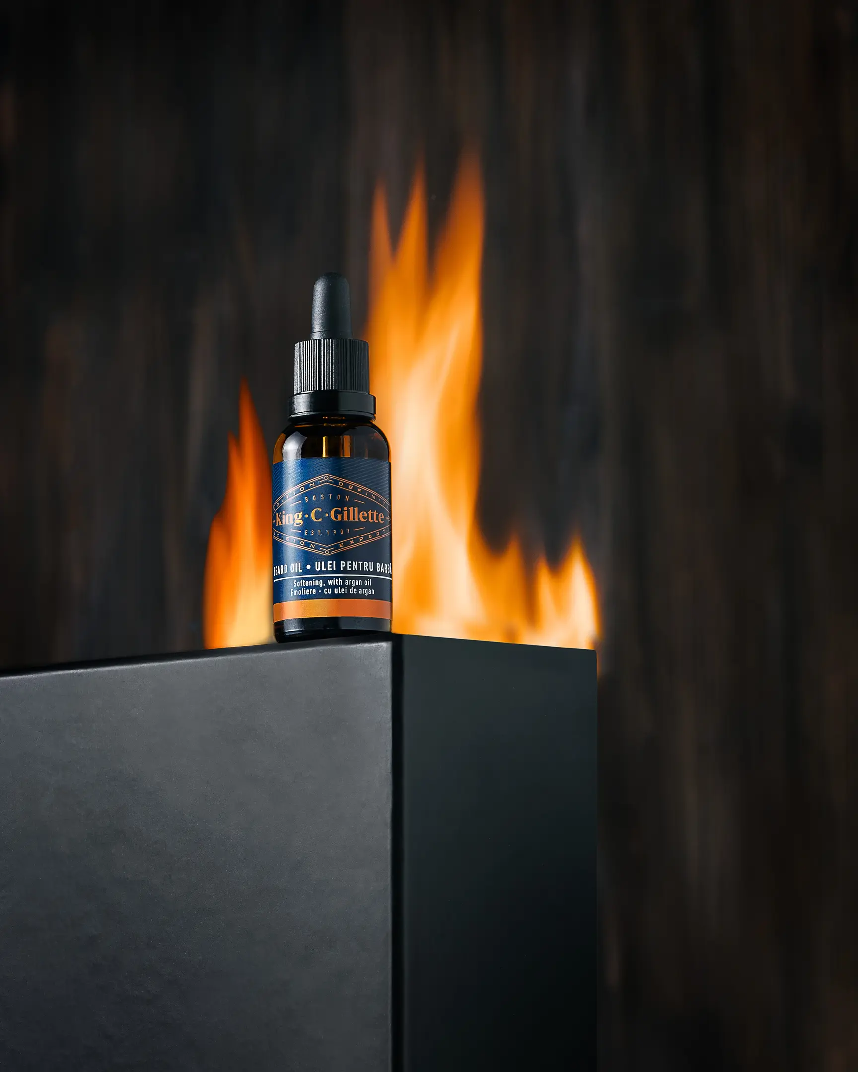 Result photo for King C Gillette with fire. Pictured is King C Gillette beard oil. It stands on a black rectangular base. Behind him, a fire is burning and a wooden background is also visible.