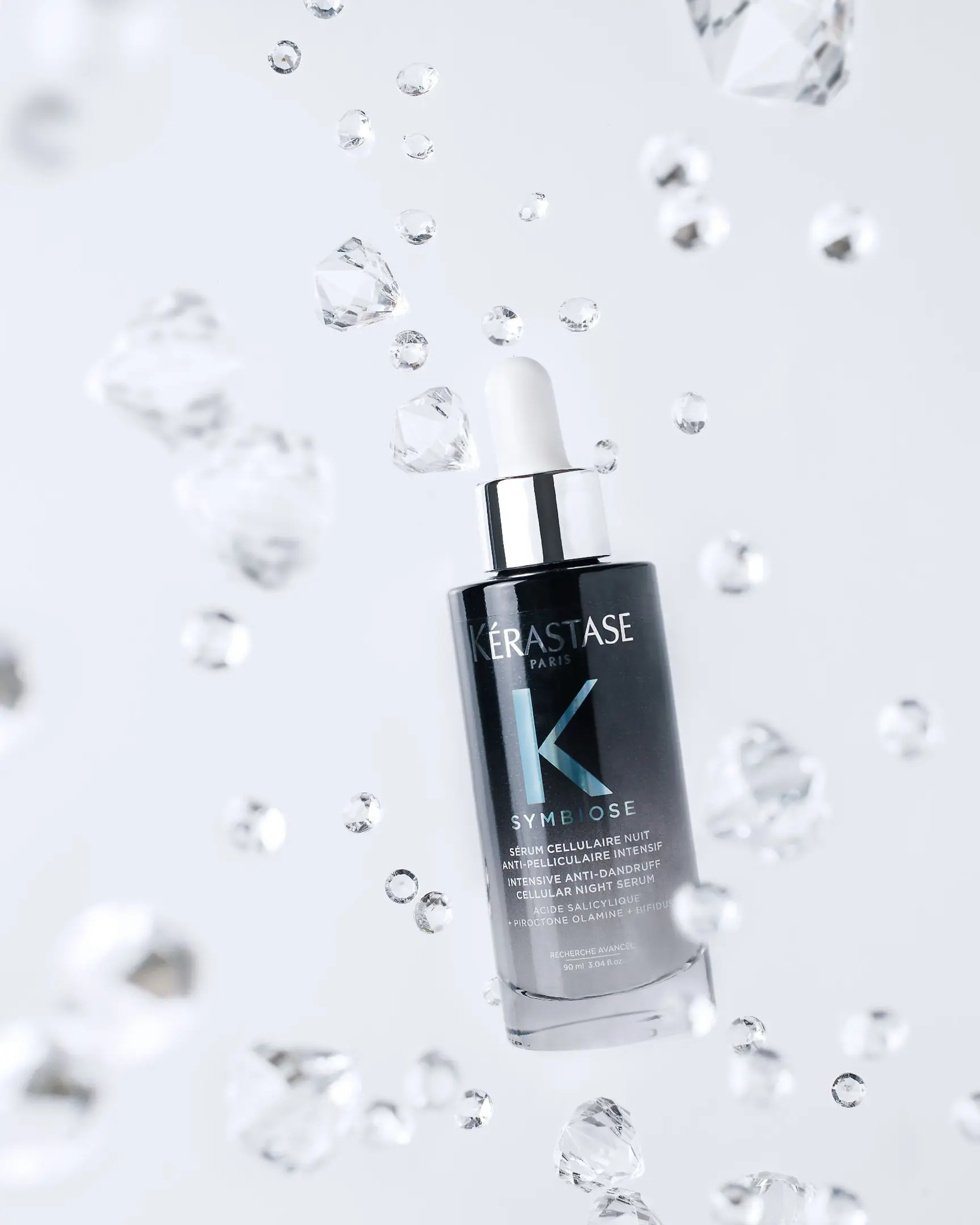 The image features a sleek bottle of Kérastase Paris "Symbiose" serum in a suspended, almost floating arrangement surrounded by crystal-like elements.  The image features a sleek bottle of Kérastase Paris "Symbiose" serum in a suspended, almost floating arrangement surrounded by crystal-like elements. The primary colors are:

    The Bottle:
        Dark grey or black, with reflective metallic accents on the label and dropper cap.
        The brand name "Kérastase" is in white, with a large blue "K" prominently featured on the bottle.

    Surrounding Crystals:
        Clear or transparent, resembling diamonds or droplets, creating a floating, luxurious aesthetic.

The background is a light grey or white, which contrasts against the darker bottle and gives the floating crystal effect a sense of depth and elegance. The overall look is minimalist, modern, and high-end.