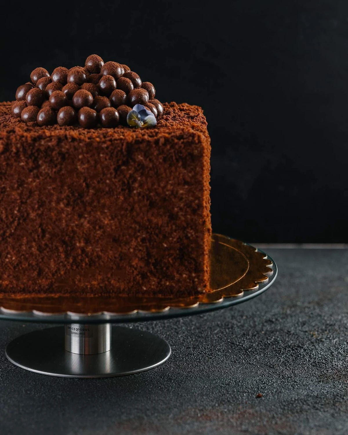 Biscuit chocolate honey cake . On a dark background, there is a square cake on a cake stand. It is decorated with chocolate balls and violet flowers.
