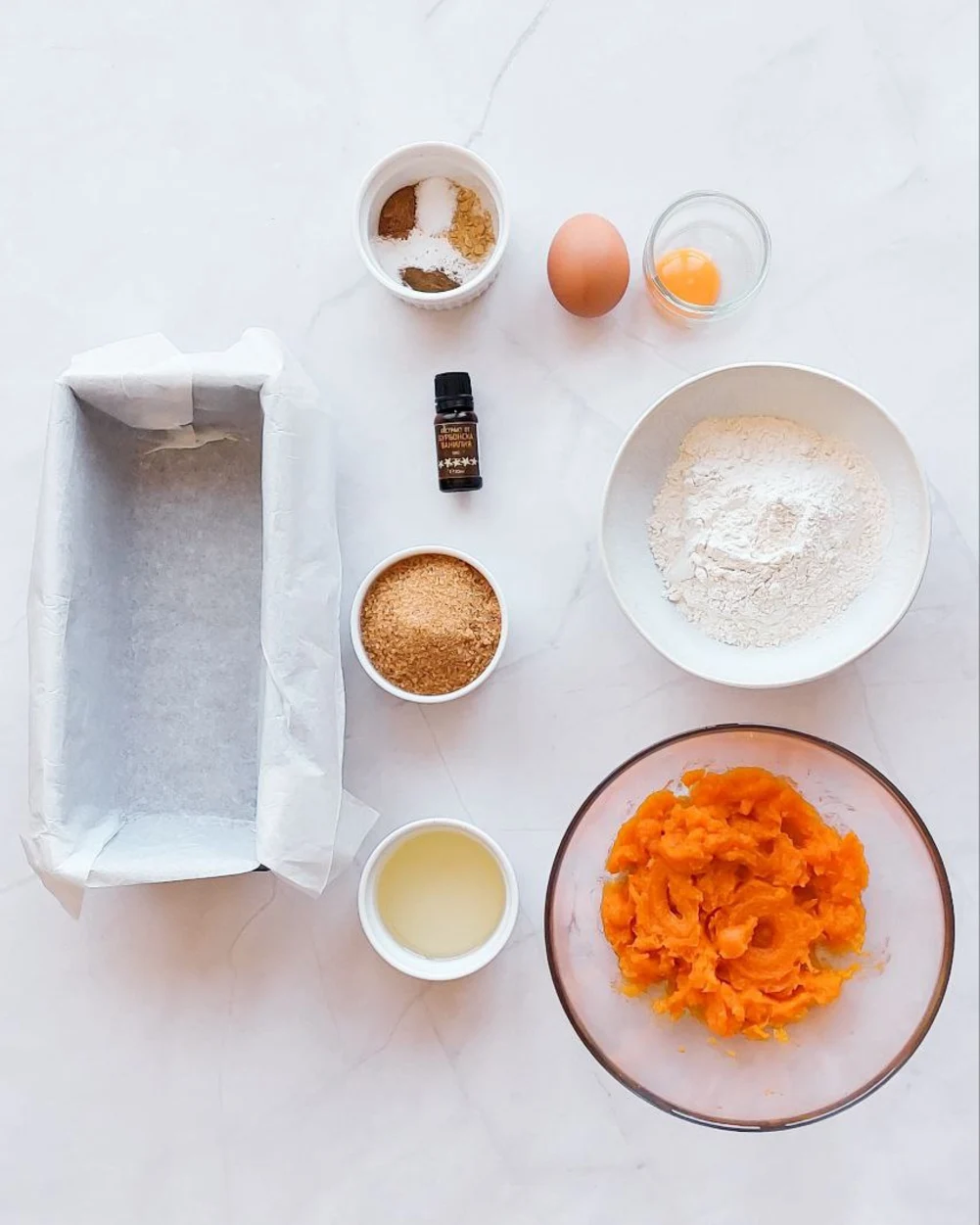 Pumpkin bread ingredients. There are several small plates on the table with different ingredients for pumpkin bread. Spices, eggs, vanilla extract, flour, and the baking dish itself.