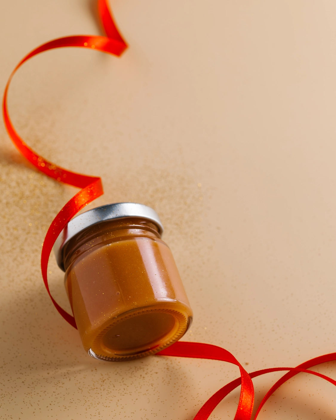A jar of caramel lies on a beige background. A jar of caramel lies on a beige background. Near it is a red ribbon, which emphasizes the presence of a festive mood and gives a reference to the celebration of the new year.