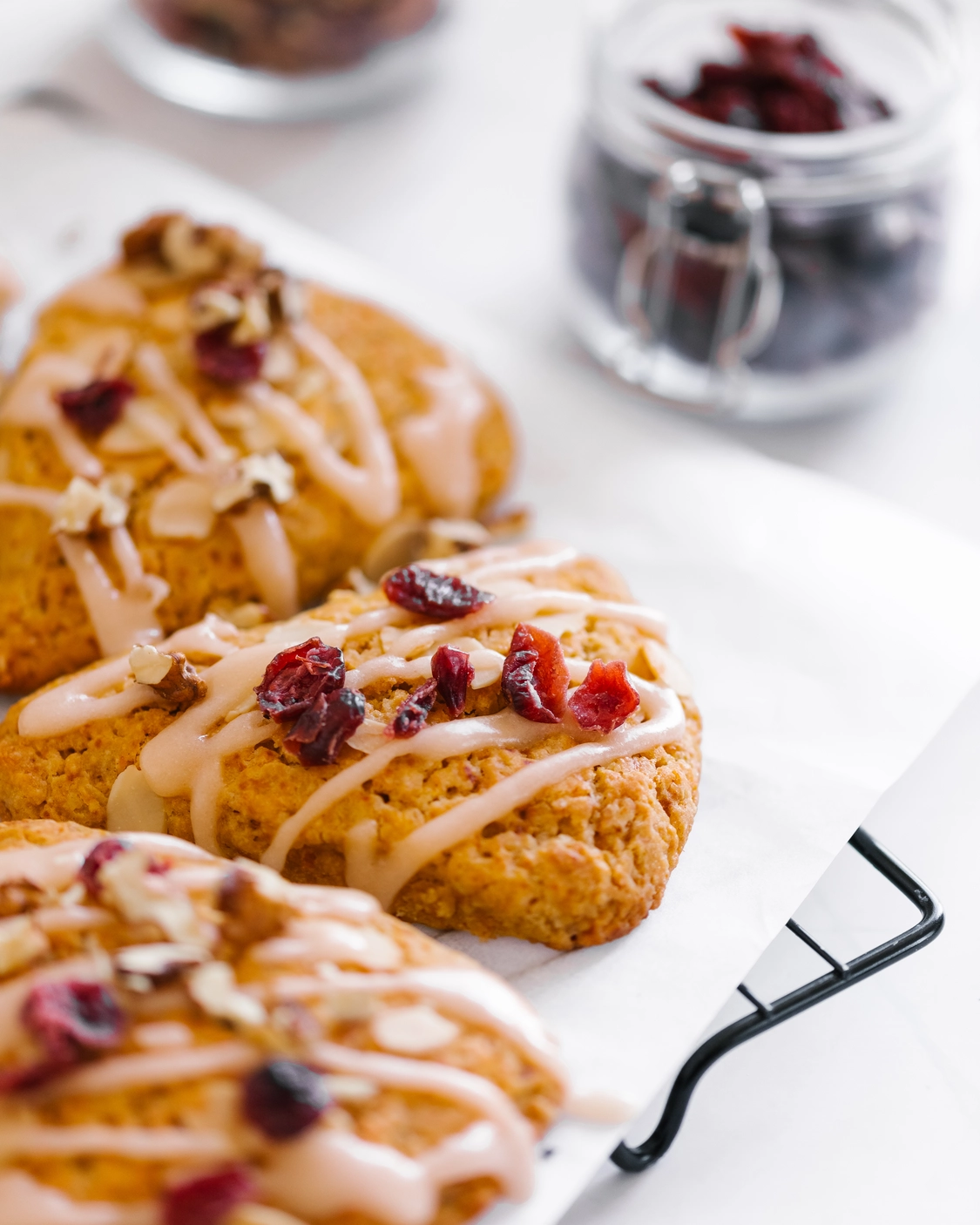 Triangles of pumpkin scones with icing. Triangles of pumpkin scones are covered with icing and dried fruits lie on baking paper on a cooling rack. Also on the table is a jar of dried cranberries - which was used to decorate the scones.