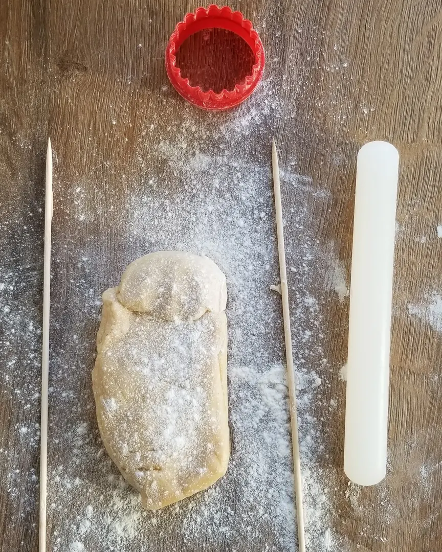 как да направите бисквитки на равномерно разстояние. Тестото лежи върху плота, поръсен с брашно, до тестото има дървени шишчета за барбекю, форма за изрязване на меденки и точилка.