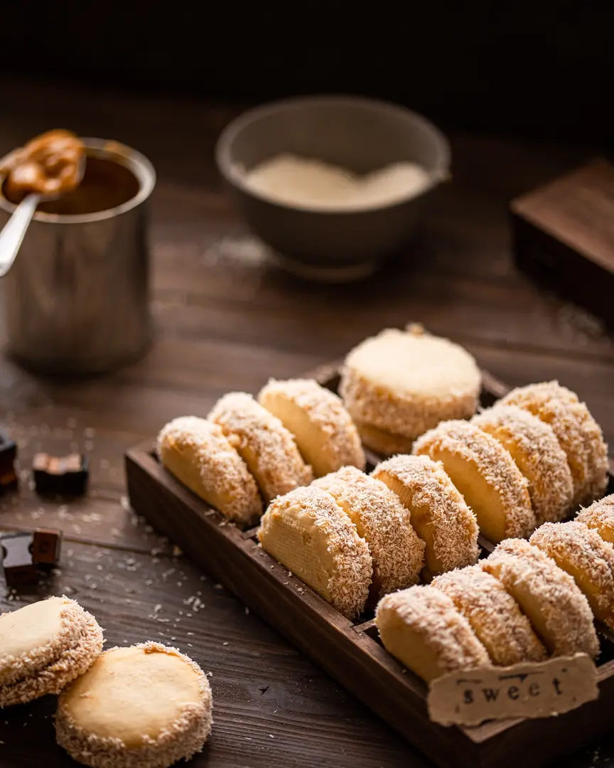 sandwich cookie filled with caramelized condensed milk.  In a square shape with cells are cookies - sandwiches with boiled condensed milk and coconut. In the foreground, there are 2 cookies on a wooden table. In the background, the viewer notices a tin can on which lies a spoon with the remains of caramelized condensed milk, and then a plate with coconut flakes.
Near the cookie is the inscription "sweet" printed on a typewriter