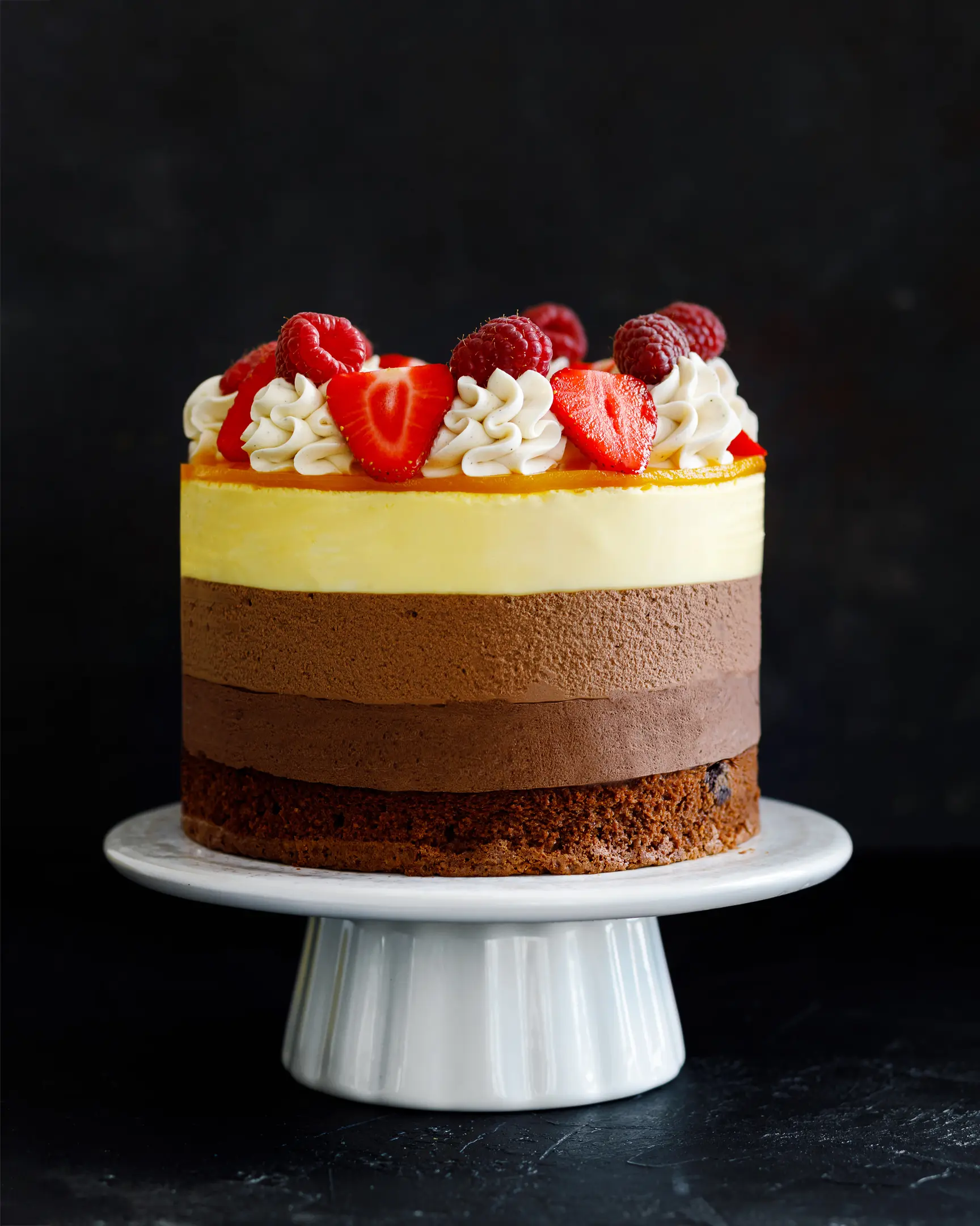 На снимката е торта от три шоколада. Пълен изглед. На снимката е торта от три шоколада. Виждат се и трите слоя с различни цветове. Тортата е украсена с малини и ягоди.