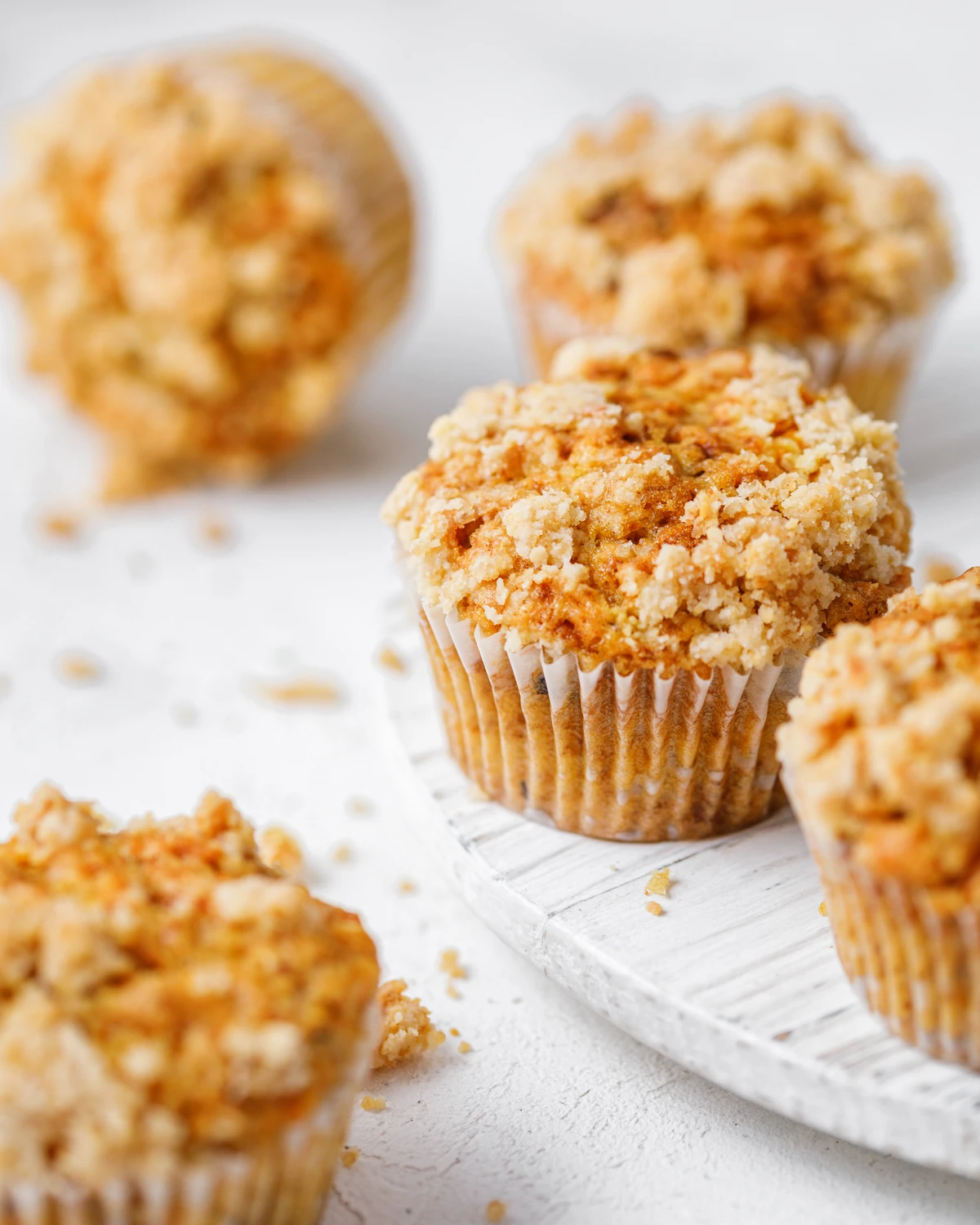 Carrot cupcakes on the table. On a light background stands a round wooden tray. In the center of the frame on a tray is a cupcake with a crispy top. Cupcakes are also on the countertop. There are some crumbs on the counter between the cupcakes.