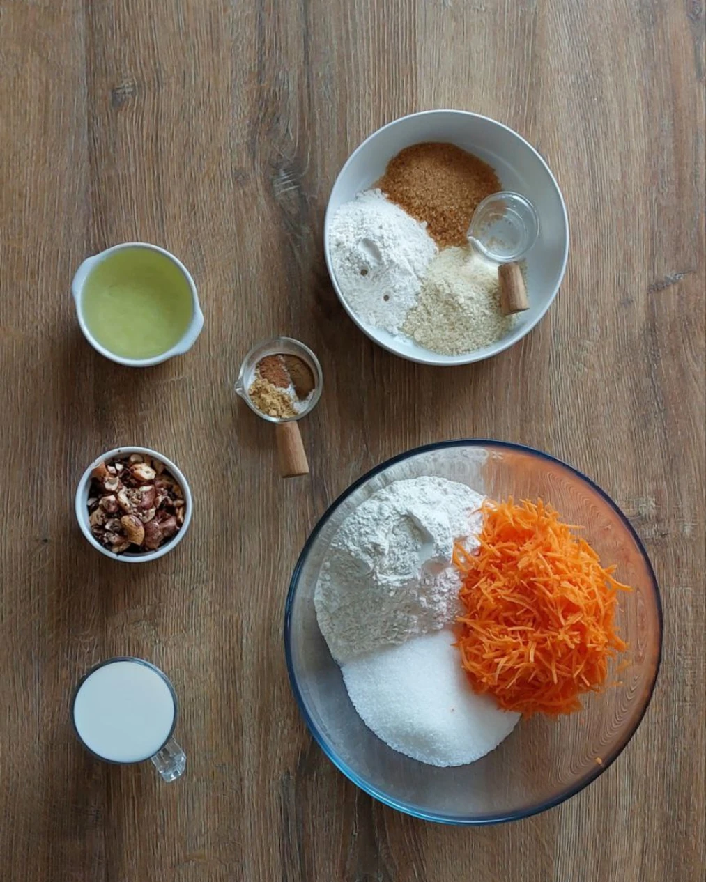 Ingredients for the carrot muffins on the table. There are muffin ingredients on the table. In a large glass container are the ingredients for the muffin - sugar, flour, and carrots. Nearby stands cups with milk, nuts, spices and vegetable oil. Separately, on top of the photo is a container with ingredients for a crispy top - nut flour, butter, flour and brown sugar.