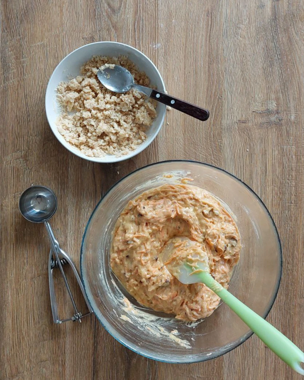 Mixed ingredients for the carrot muffins on the table. Mixed ingredients for muffins are on the table. Muffin batter is placed in a large glass container. On a small plate - mixed the crumble. Separately, there is a spoon for ice cream - so that it is more convenient to make cupcakes of the same size.