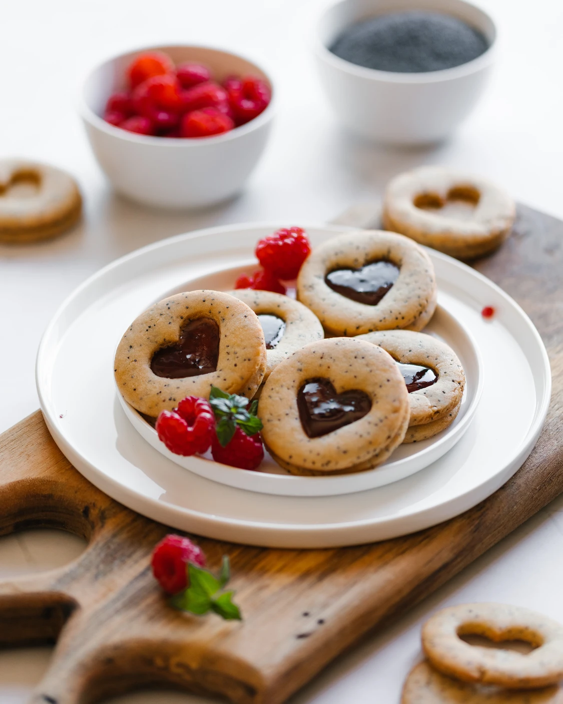 Vegan poppyseed cookies are on the table. On a wooden cutting board, there is a ceramic plate on which cookies with poppy seeds lie. The cookies are sandwiched in the middle with jam.
In the background of the picture are plates with raspberries (from which the jam is made) and poppy seeds.