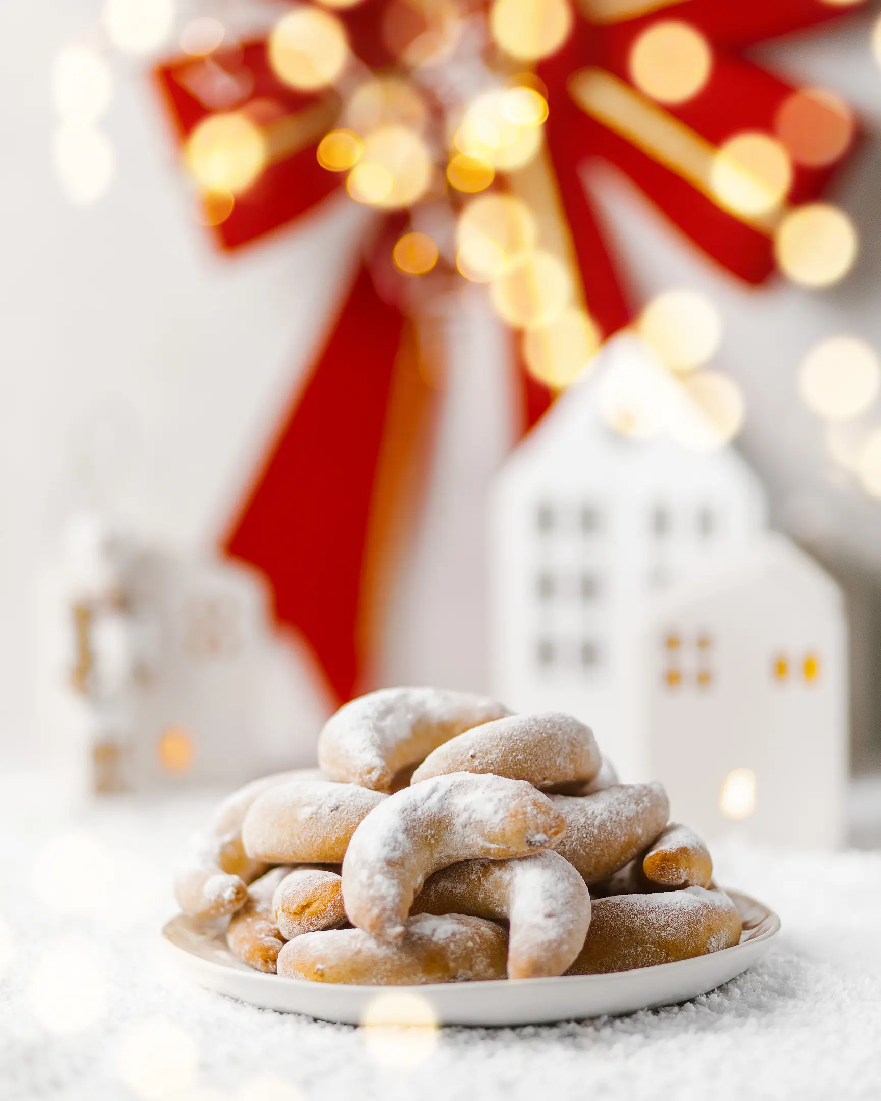Сrescent-shaped cookies. On the table is a plate of crescent-shaped cookies. A red bow hangs on the wall in the background. Beautiful bokeh from the Christmas tree garland is visible in the frame. Next to the bow are ceramic houses.