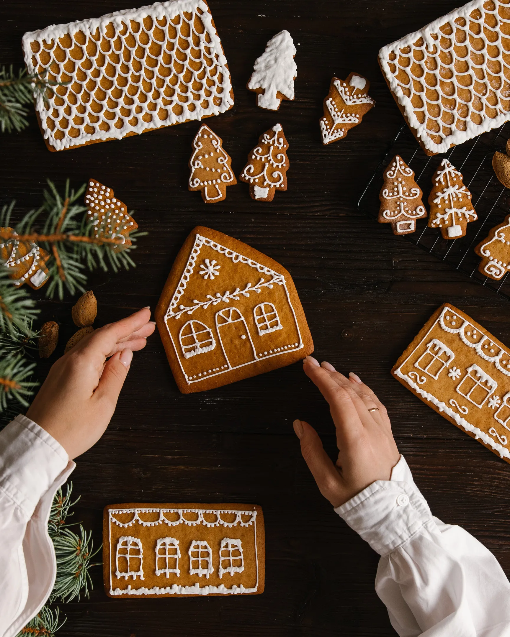 On the table are gingerbread in the form of walls and the roof of the house On the table is gingerbread in the form of walls and the house's roof. A woman is holding 1 side of the house. To the right of the frame are almond nuts. Christmas tree needles are visible in the foreground.