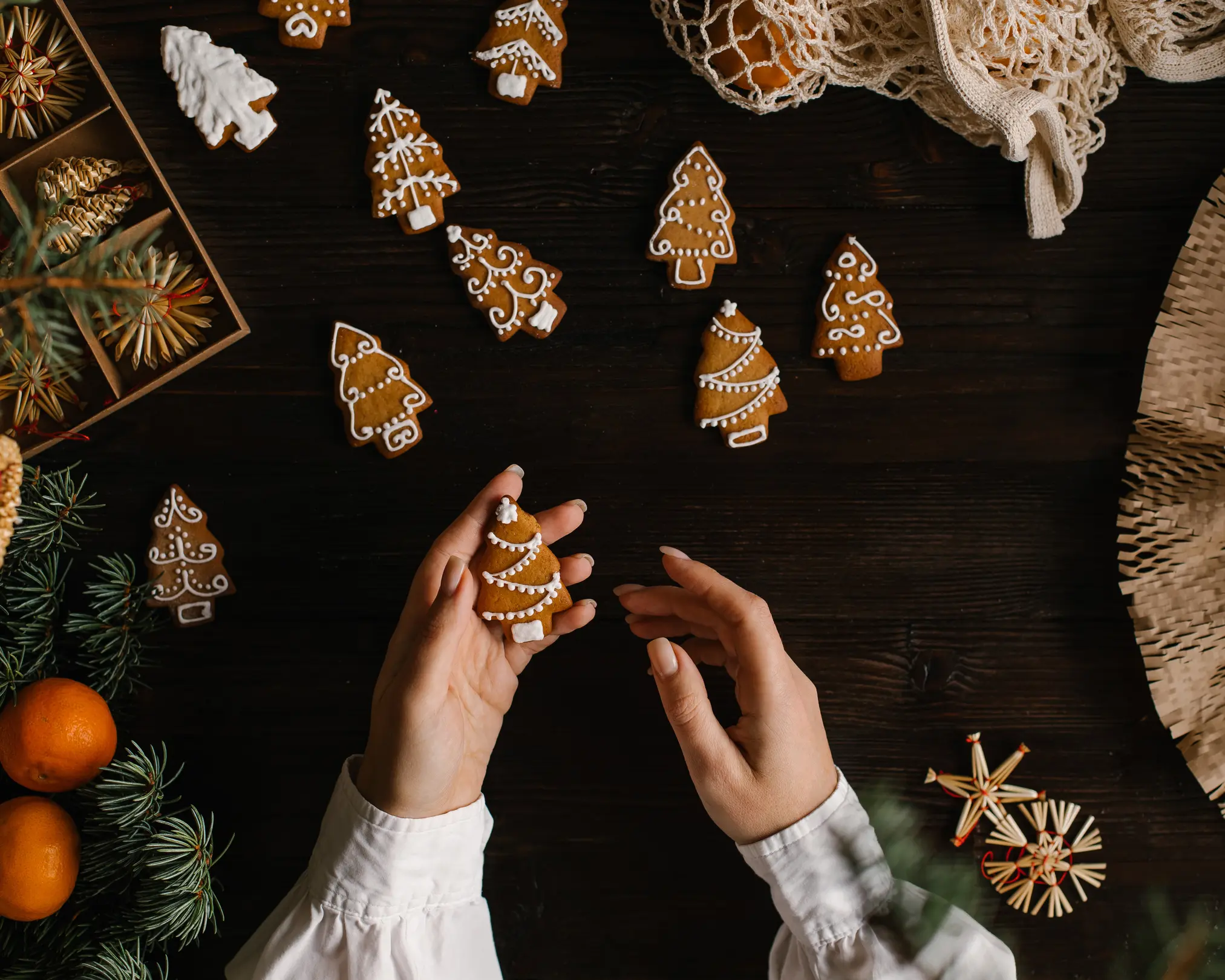 On the table is gingerbread in the form of trees. On the table is gingerbread in the form of trees. A woman is holding 1 side of the cookies. To the right of the frame are almond nuts. Christmas tree needles are visible in the foreground.