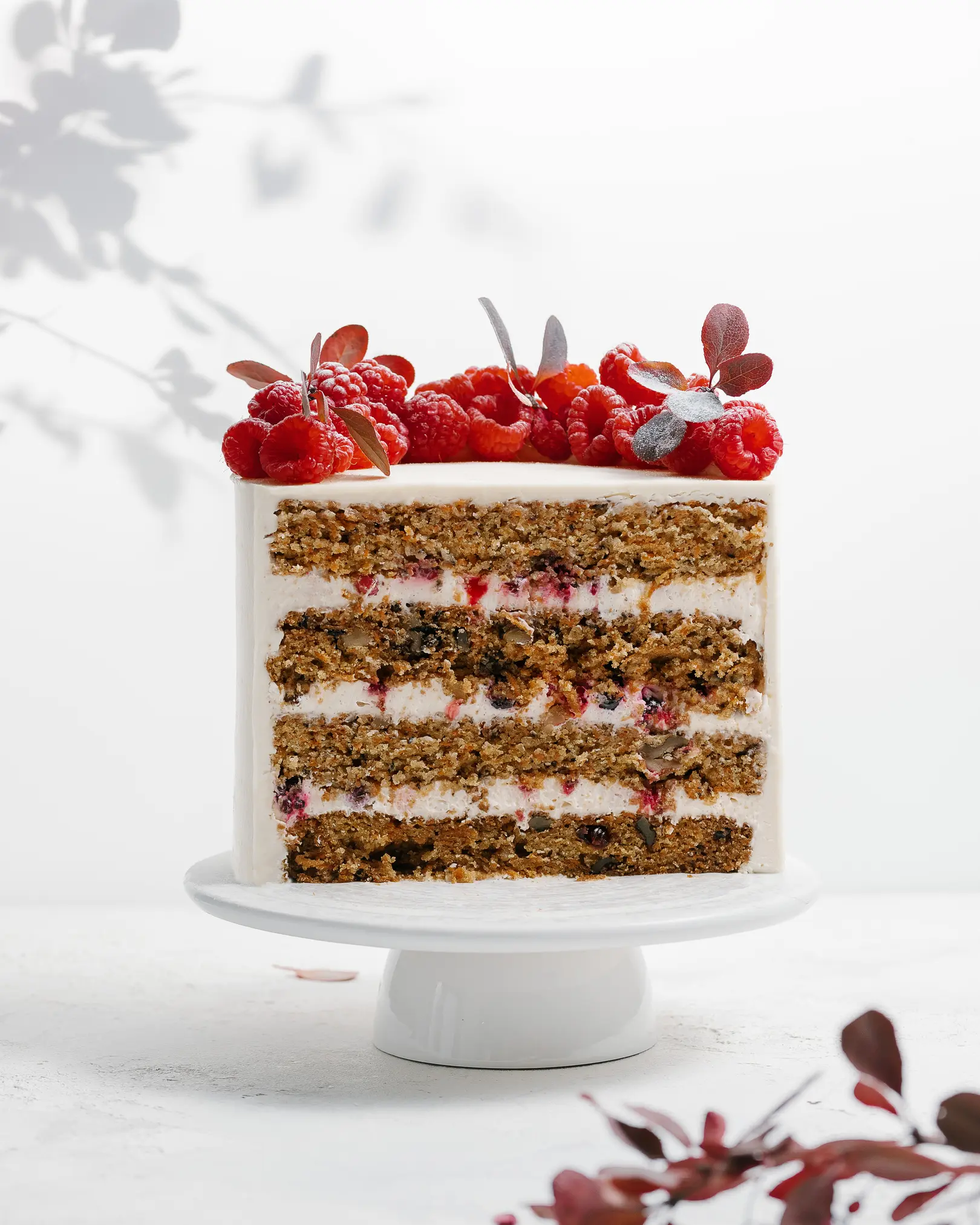 Carrot cake with berries into the layer. There is a cake on the table. It is decorated with raspberries. Visible carrot cakes, creamy layers, and chocolate cream cover the outside of carrot cakes. In the foreground lies a sprig of a plant with red leaves, the same leaves appear through the background.