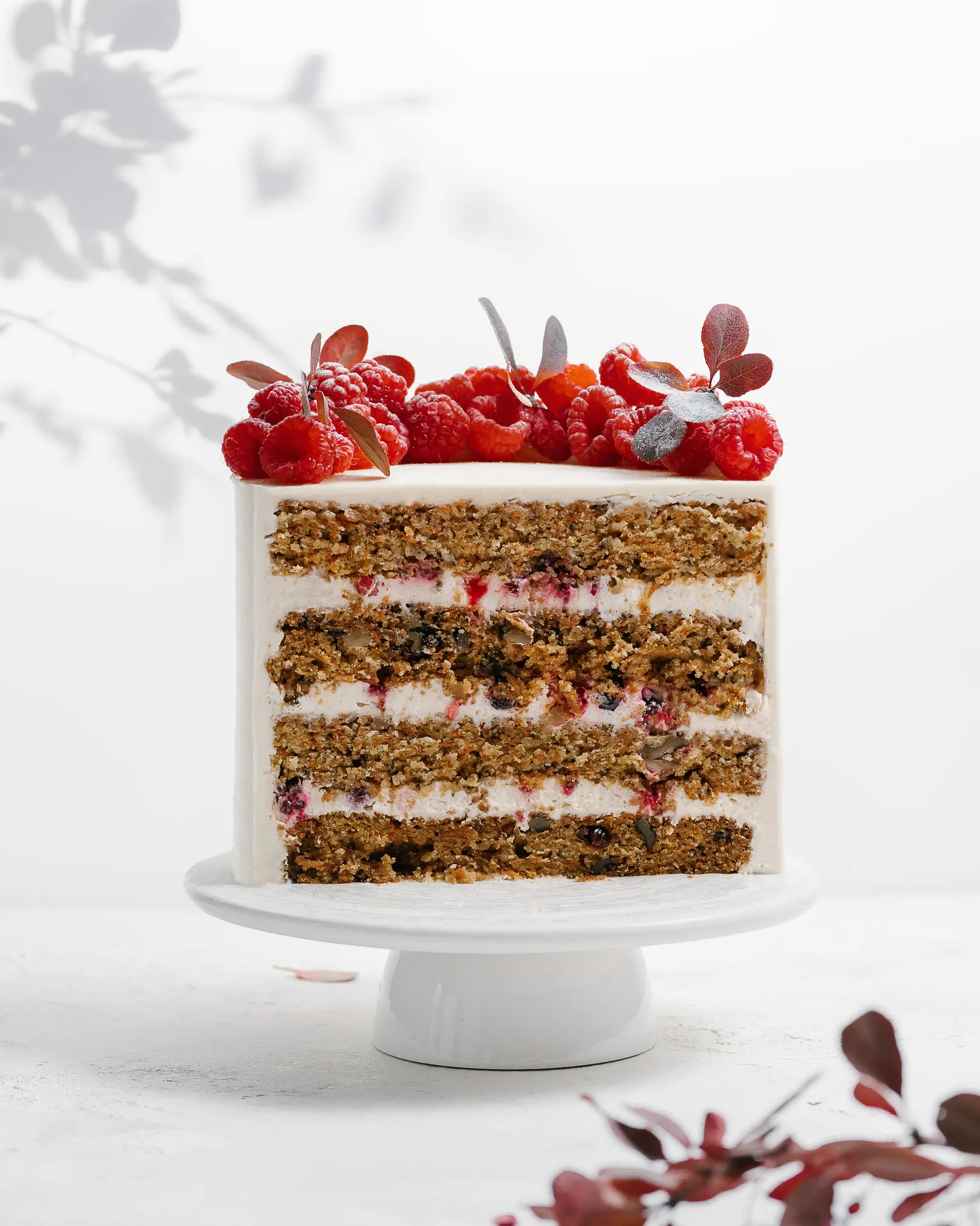 There is a carrot cake.  There is a cake on the table. It is decorated with raspberries. Visible carrot cakes, creamy layers, and chocolate cream cover the outside of carrot cakes. In the foreground lies a sprig of a plant with red leaves, the same leaves appear through the background.