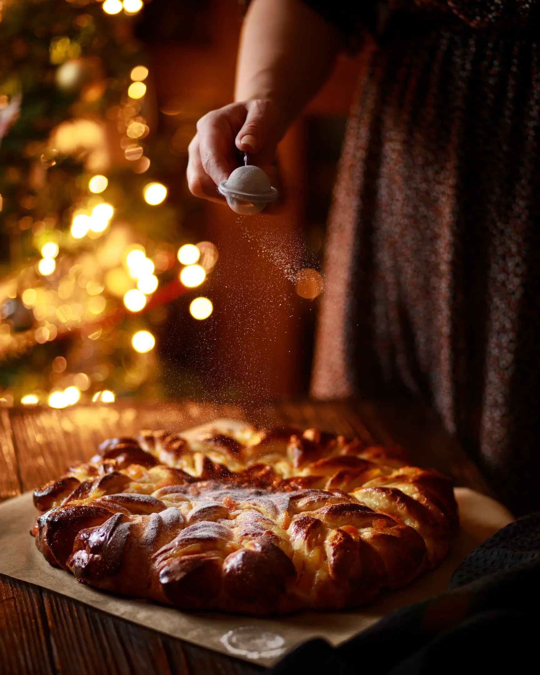On it is a star-shaped cake Against the background of flickering lights from the Christmas tree is a table. On it is a star-shaped cake. A woman sprinkles powdered sugar on top of a cake.