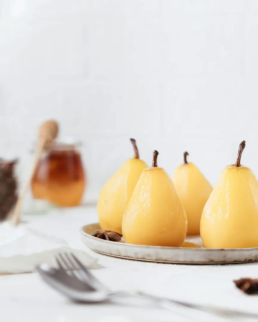 Pears poached in wine. There are light pears on a light plate. In front of them are a fork and a spoon. A jar of honey is visible in the background of the picture. Pears shine - because they are poured with sweet syrup