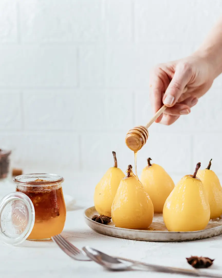 Poached pears with honey. There are light pears on a light plate. In front of them are a fork and a spoon. A jar of honey is visible in the background of the picture. Pears shine - because they are poured with honey. At the top of the picture is a woman's hand holding a honey spoon. Honey drips from a spoon onto pears.