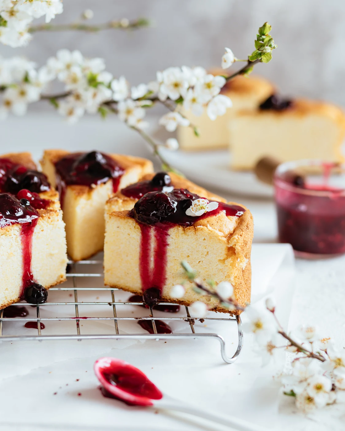 Russian cottage cheese pie on a table. On the table, on a lattice under flowering cherry branches, there are slices of cottage cheese casserole. Cherry sauce on top of the casserole. It drains. In the foreground lies a spoon smeared in cherry sauce.