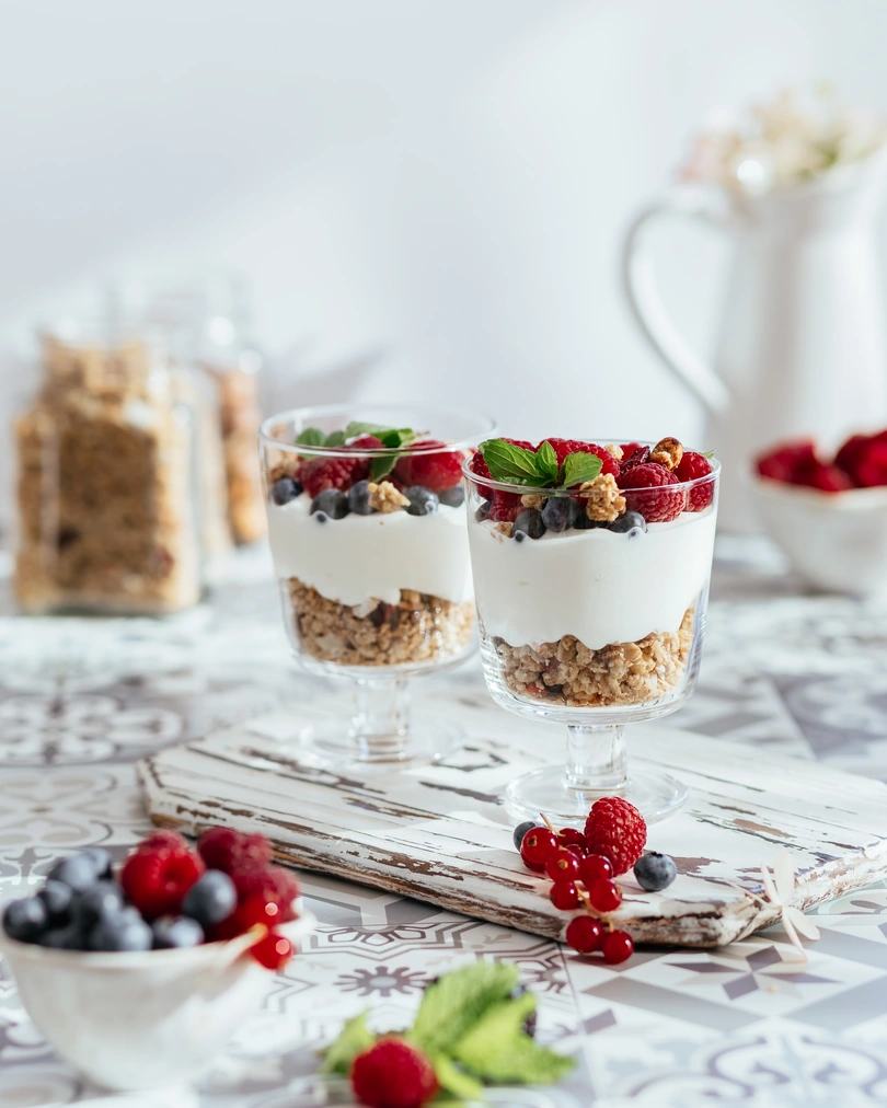 2 cups filled with granola and berries. 2 cups filled with granola. On top of the granola is a layer of yogurt. There are many berries on top. The cups are placed on a wooden stand. Various berries are visible in the foreground. In the background of the photo are a jar of granola and a milk jug.