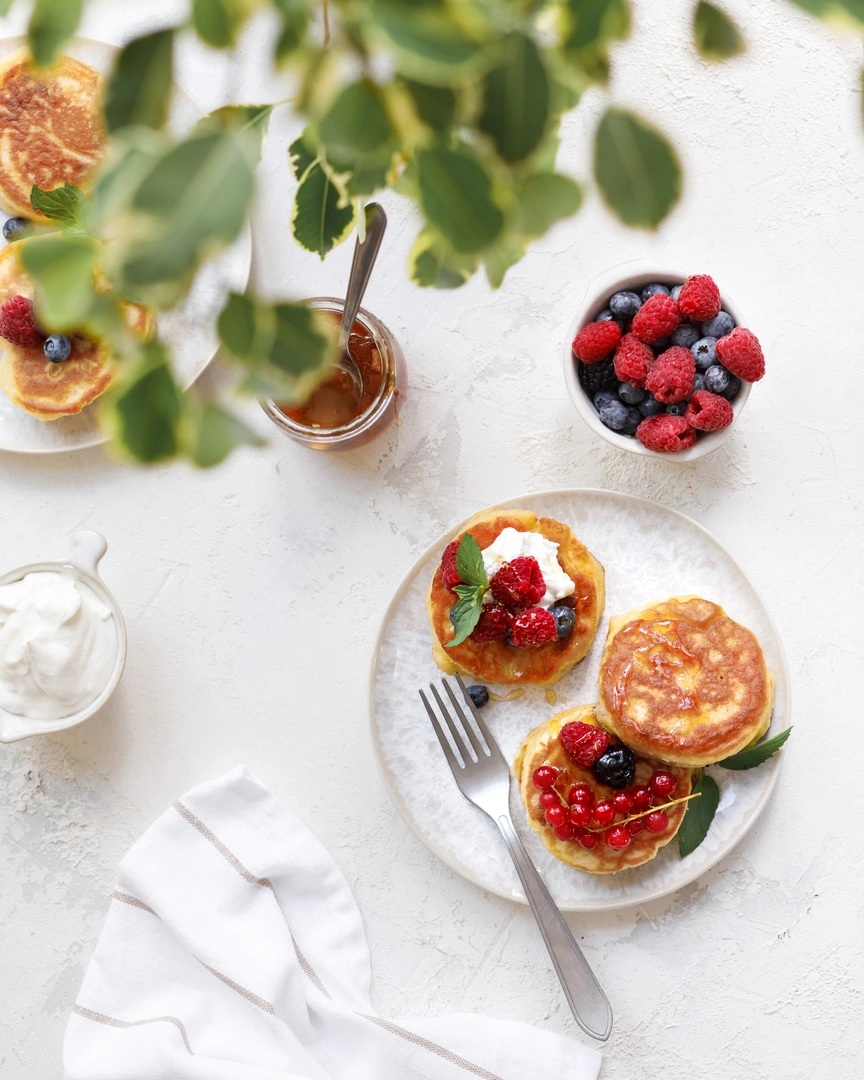 Lush pancakes. There are several plates on a light table. On one of the plates are fluffy pancakes. On the plate lie berries covered with honey. To the left of the plate is a jar of sour cream.