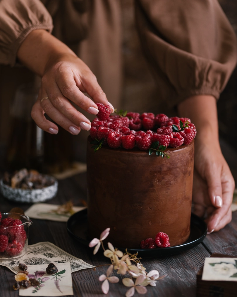 On the table is a cake "Milk Girl". A girl in a brown dress decorates a cake covered with chocolate with raspberries. In the foreground lies caramelized sugar. In the blur, raspberries are visible on a transparent plate.