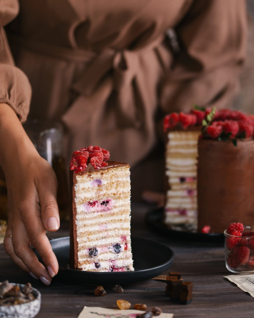 На тъмна дървена маса парче торта. Момиче в кафява рокля украсява торта, покрита с шоколад и малини. На преден план е карамелизирана захар. В замъгленото изображение се виждат малини в прозрачен контейнер.
