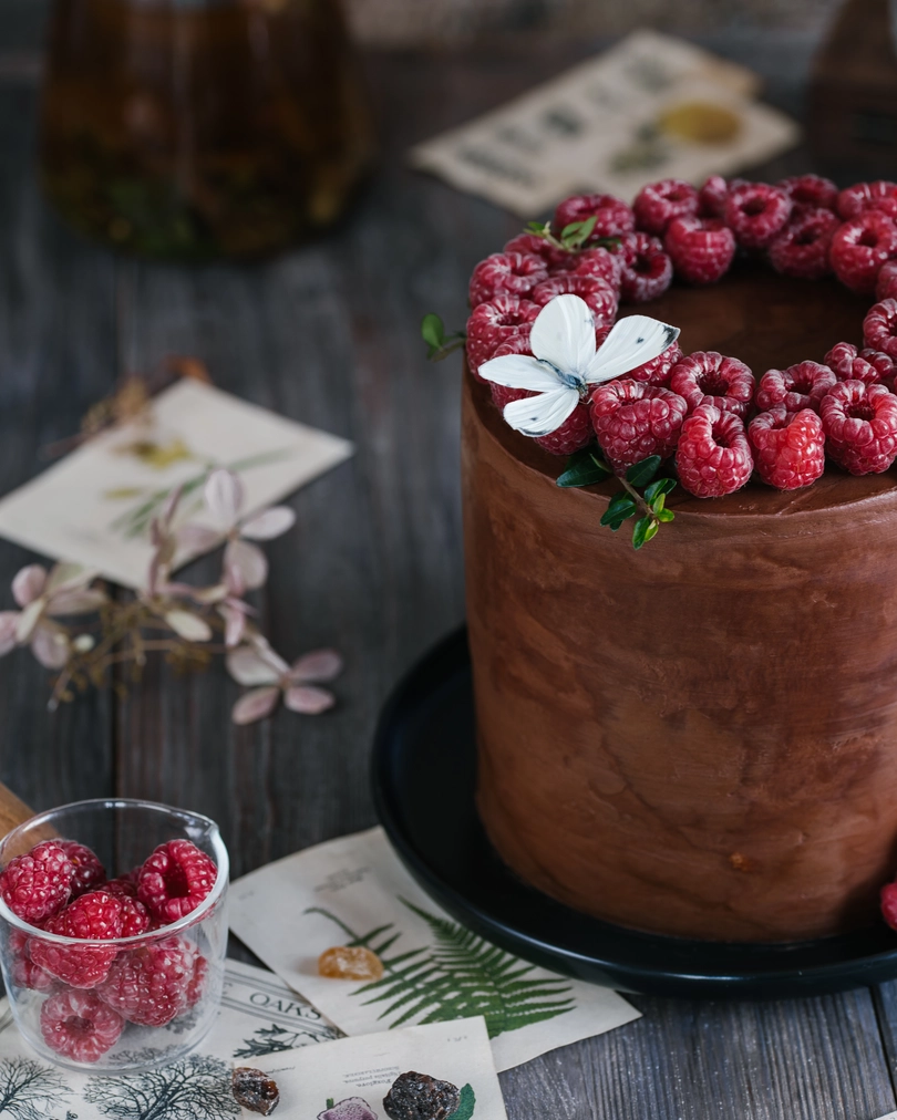 On the table is a piece of cake "Milk Girl" with butterfly on the top. On the table is a cake. It is covered with chocolate and raspberries. In the foreground lies caramelized sugar and old postcards. A butterfly landed on top of the cake