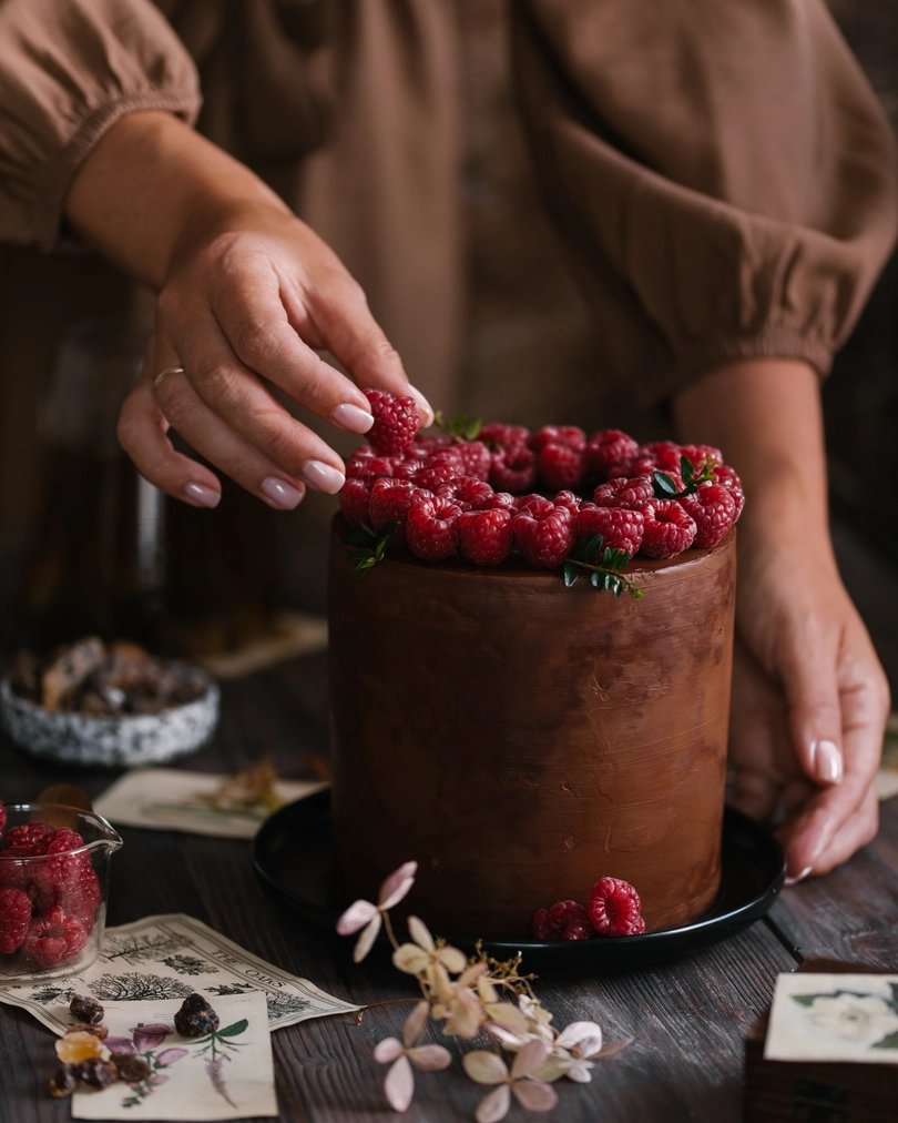 On the dark table is cake with raspberries. A girl in a brown dress decorates a cake covered with chocolate with raspberries. In the foreground lies caramelized sugar. In the blur, raspberries are visible on a transparent plate.