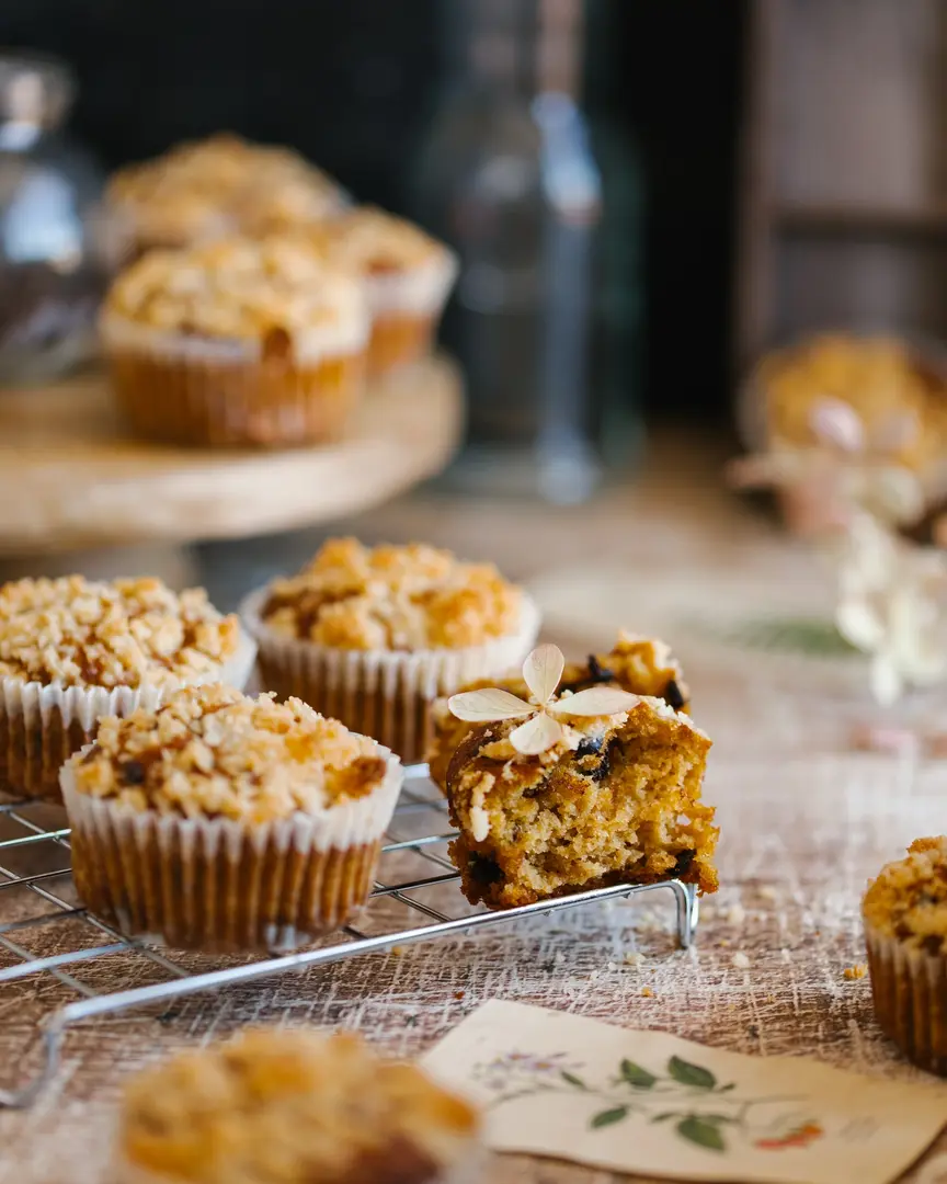 Pumpkin muffins. In the foreground of the photo - on the surface of red color with scratches lies a paper card with flowers, in a blur in the foreground cupcakes. The viewer's gaze moves from the cupcakes to the postcard and onto the broken cupcake. It has chocolate chips in it. In the background, on a wooden cake stand, there are the same cupcakes. And the blurred wooden shape in the upper right corner completes the composition.